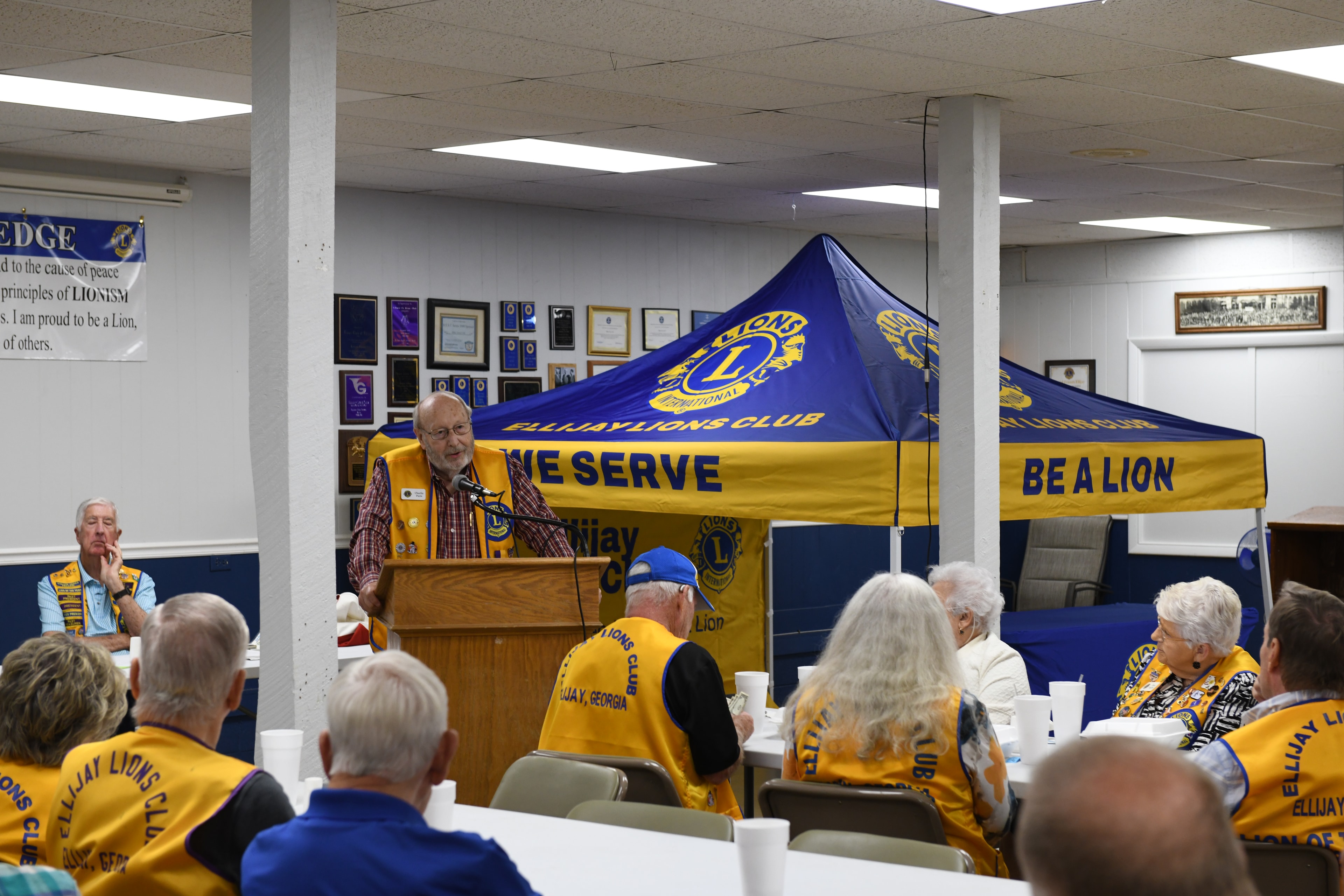 Lions Club member speaking at the podium during a monthly meeting with members seated